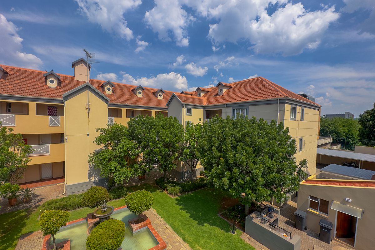 An elevated aerial view of an apartment complex featuring a well-maintained communal garden, central water feature, and terracotta-tiled roof structures.