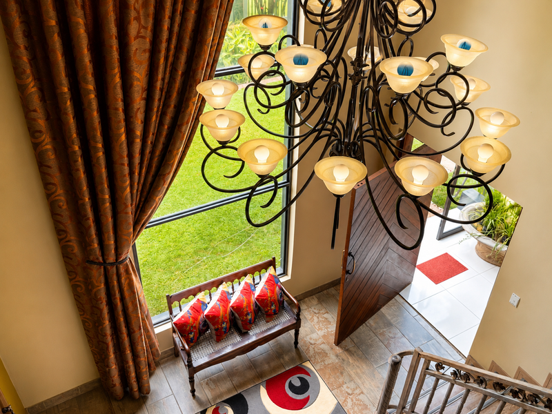 A grand entrance hall featuring a double-height window, a large decorative chandelier, tiled flooring, and a prominent wooden pivot door.
