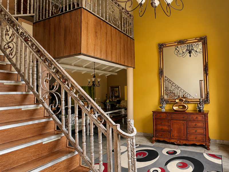 Spacious entrance hall featuring a grand staircase with ornate wrought iron railings, wood-paneled mezzanine, and high ceilings with a decorative chandelier.