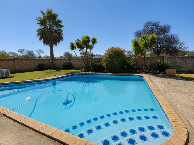 A well-maintained outdoor swimming pool area featuring a paved surround, lawn, and established garden landscaping enclosed by a brick boundary wall.