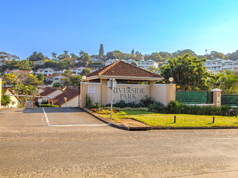 The entrance to Riverside Park retirement village, featuring a secure gated access point, paved driveway, and well-maintained landscaped gardens.
