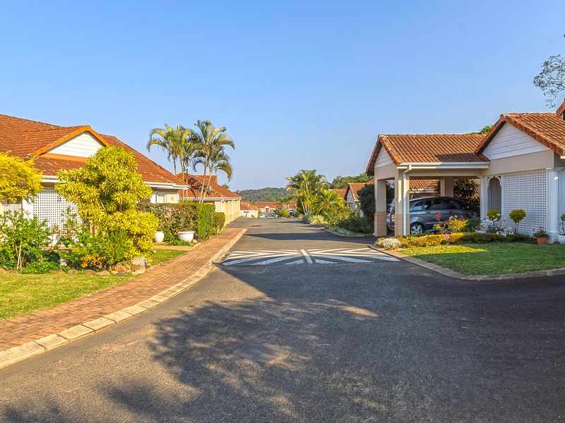 A view of a well-maintained retirement village street featuring paved driveways, carports, and established landscaped gardens under a clear blue sky.