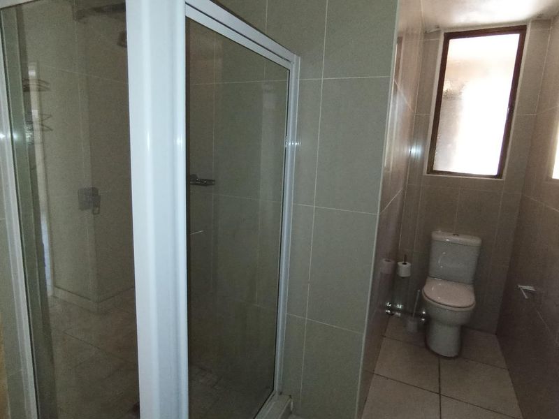 A functional bathroom featuring a glass shower enclosure, a standard toilet, and neutral wall and floor tiling with a frosted window for natural light.