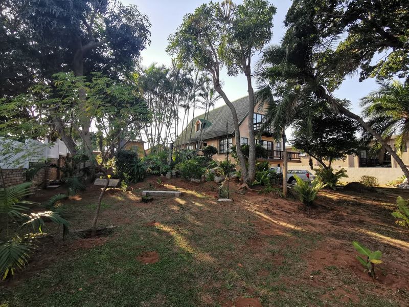 A view of the rear garden featuring mature trees, established landscaping, and a multi-story house with a balcony.