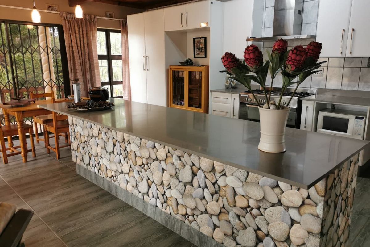 Spacious kitchen featuring a unique stone-clad island with a stainless steel countertop, built-in appliances, and timber ceiling beams, opening to an outdoor area.