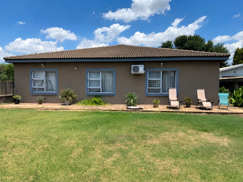 Rear view of the house featuring a paved patio area, louvered windows, and a wall-mounted air conditioning unit overlooking a spacious lawn.