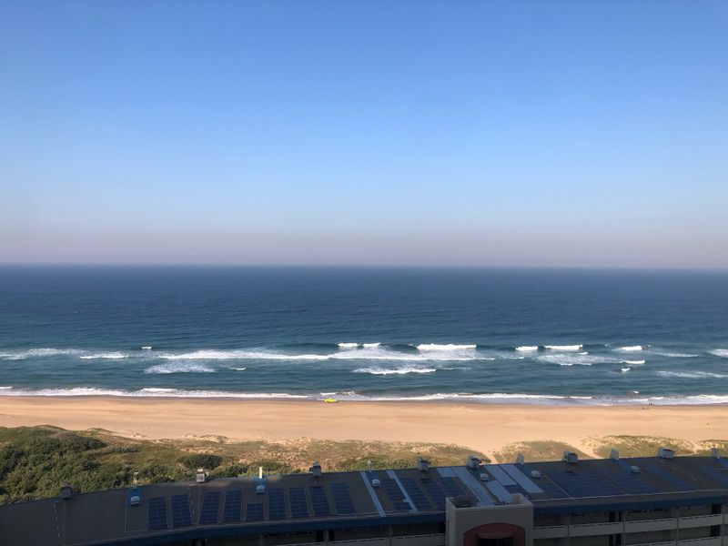 A clear, elevated view of the ocean and sandy beach, featuring a building roof with installed solar panels in the foreground.