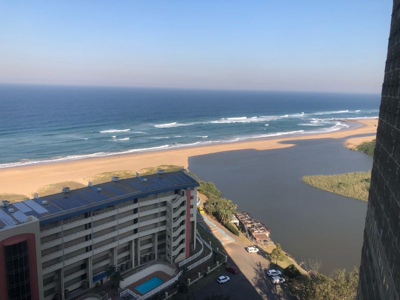 Elevated coastal view overlooking the ocean, beach, and estuary, featuring a nearby apartment complex with rooftop solar panels.