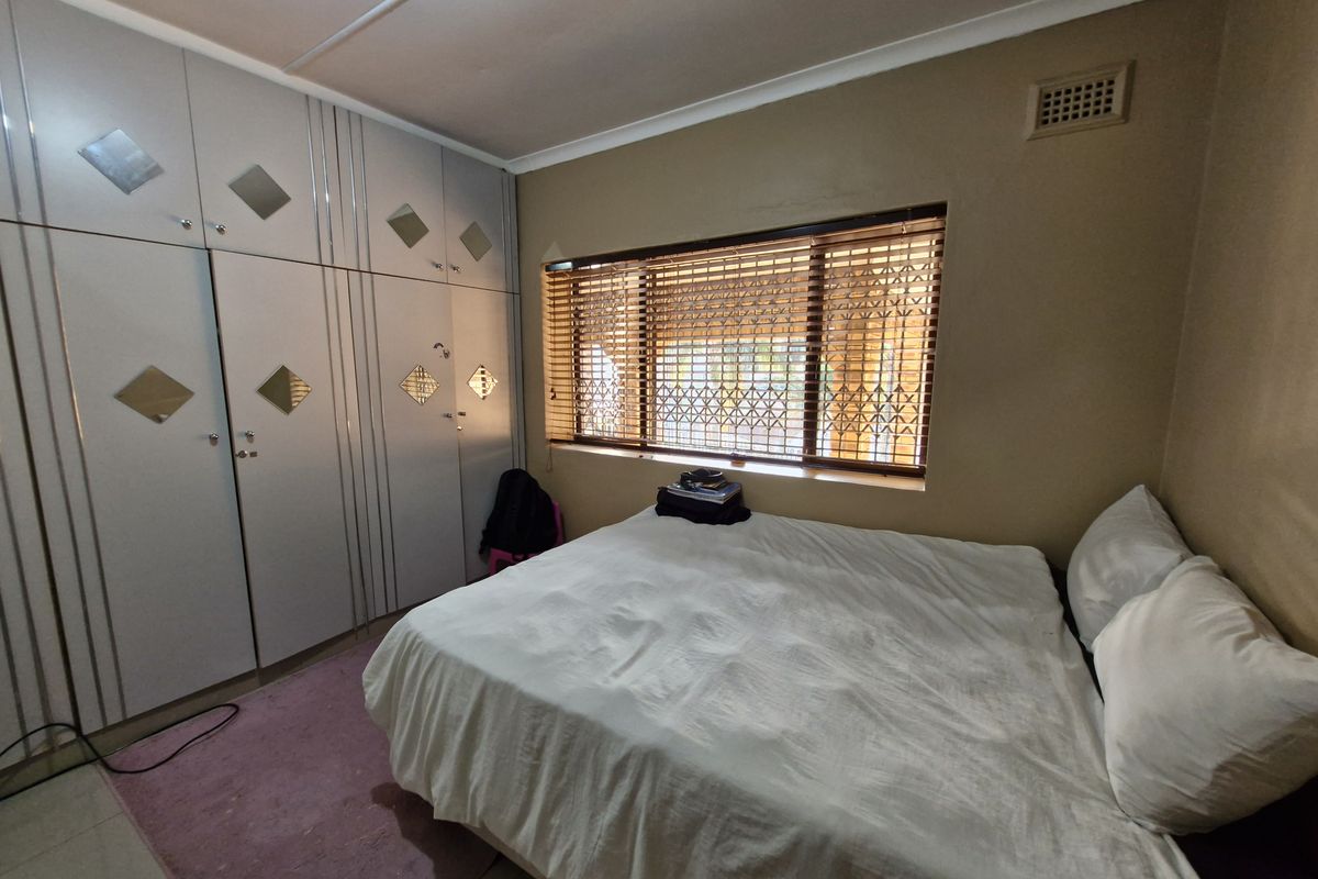 A bedroom featuring built-in cupboards, tiled flooring, and a window fitted with wooden venetian blinds and a security gate.