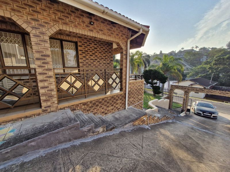 Front exterior view featuring a face brick facade, a covered patio with metal balustrades, and a concrete driveway leading to the property entrance.