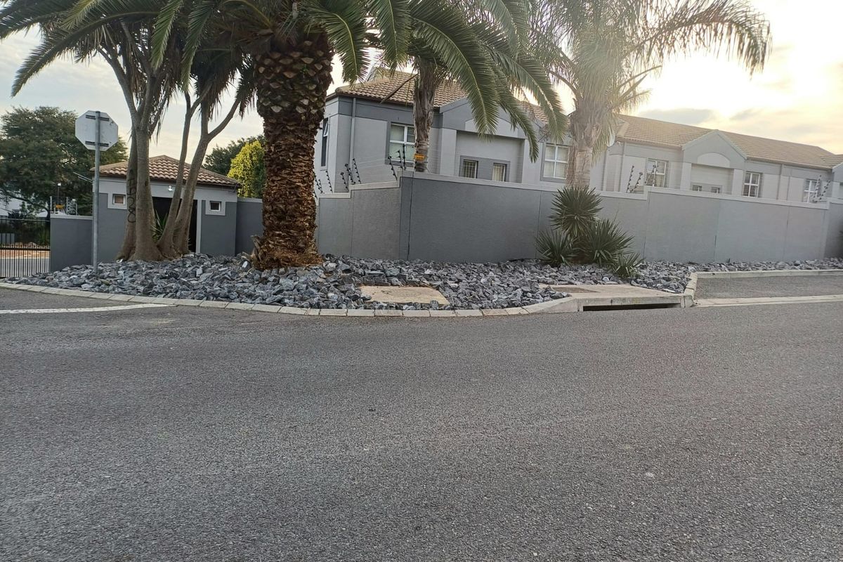 Street view of the townhouse complex exterior, featuring a perimeter wall with electric fencing, mature palm trees, and paved curb infrastructure.