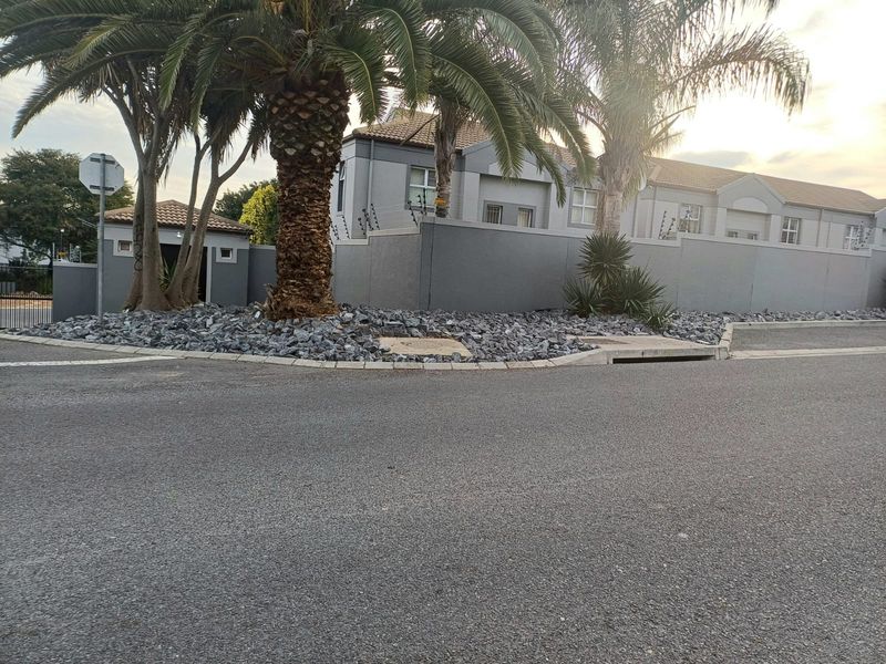 Street view of the townhouse complex exterior, featuring a perimeter wall with electric fencing, mature palm trees, and paved curb infrastructure.