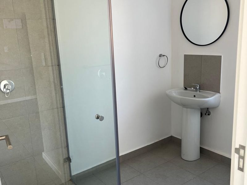 A clean and modern bathroom featuring a pedestal basin, a glass-enclosed shower with neutral tiling, and a minimalist circular mirror.