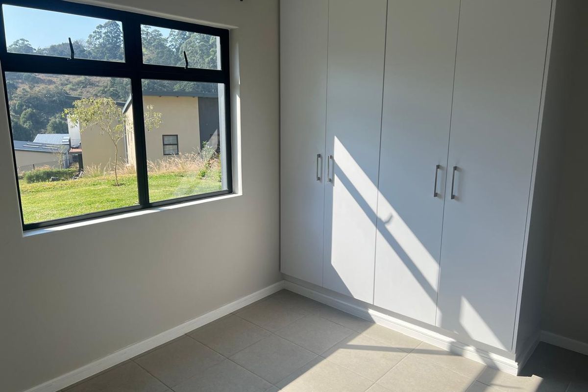 A bright, empty bedroom featuring modern built-in cupboards, neutral tiled flooring, and a large window with views of the surrounding landscape.