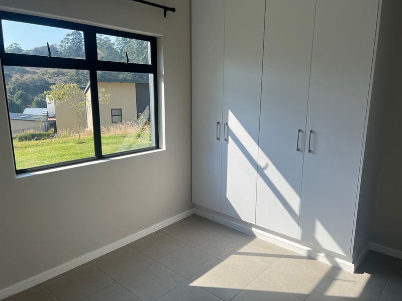 A bright, empty bedroom featuring modern built-in cupboards, neutral tiled flooring, and a large window with views of the surrounding landscape.