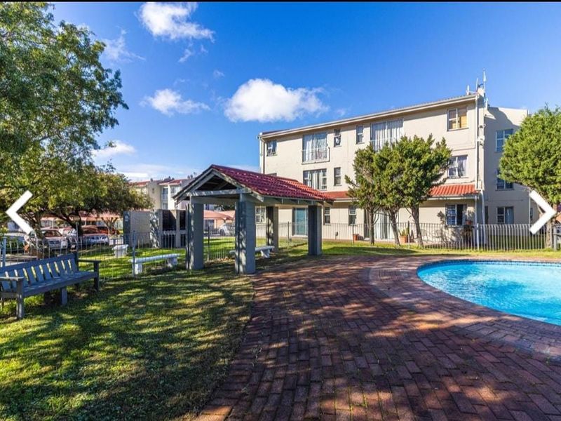 Communal pool area featuring a brick-paved deck, covered gazebo with built-in braai facilities, and landscaped garden surroundings within the apartment complex.