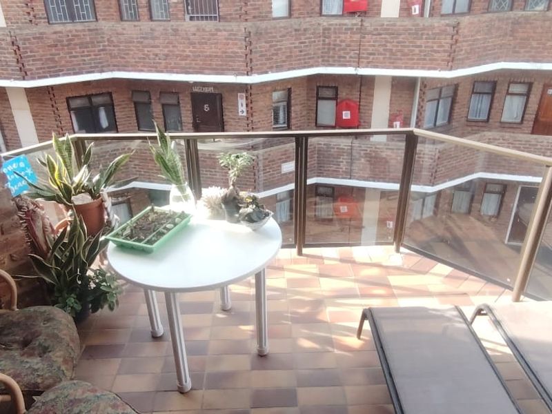 A covered balcony featuring a built-in braai, glass balustrade, and tiled flooring, overlooking the townhouse complex courtyard.