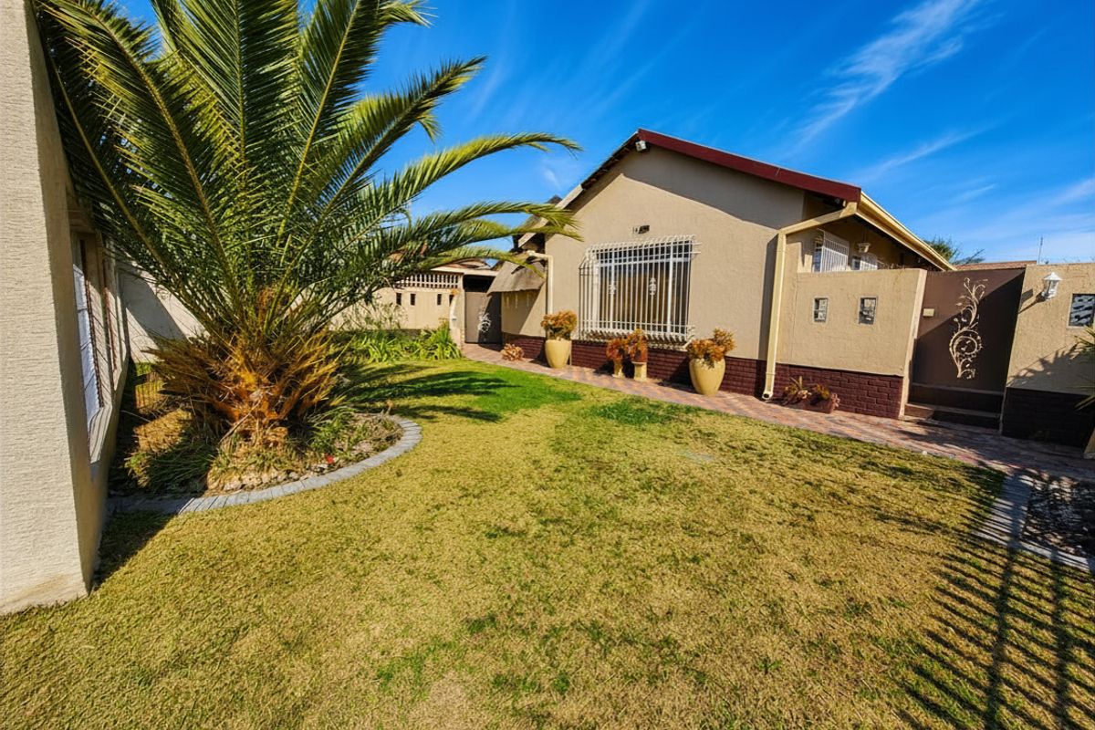 A well-maintained rear garden area featuring a lawn, mature palm tree, paved pathway, and secure exterior facade with decorative metal gate.