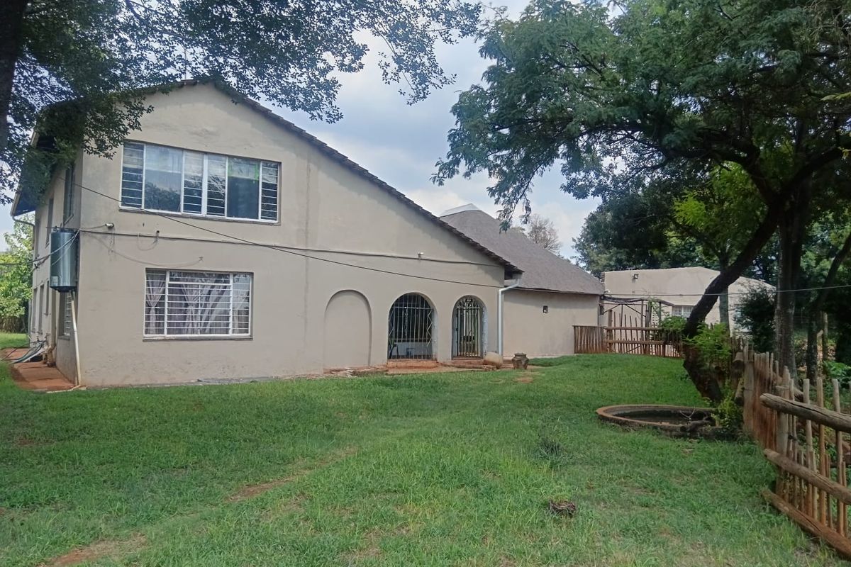 Rear view of a two-story house featuring a large garden, mature trees, arched doorways with security bars, and a tiled roof.