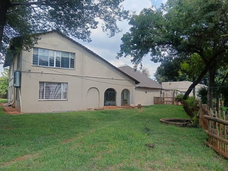 Rear view of a two-story house featuring a large garden, mature trees, arched doorways with security bars, and a tiled roof.