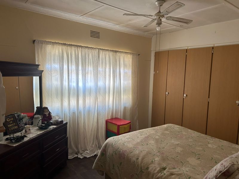 A bedroom featuring built-in wooden cupboards, a ceiling fan, and a ventilation grille. The room shows signs of wear, particularly on the ceiling.