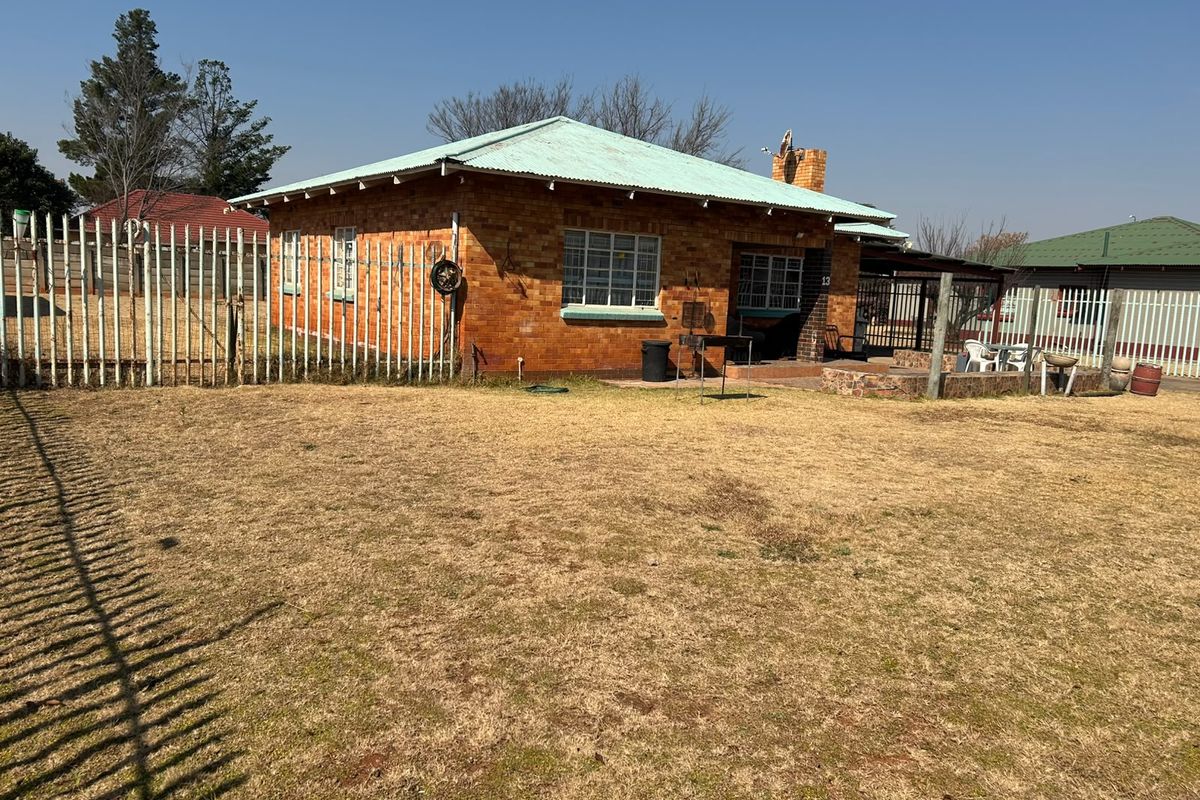 Rear view of a single-story brick house featuring a corrugated metal roof, chimney, and a paved patio area set within a fenced yard.