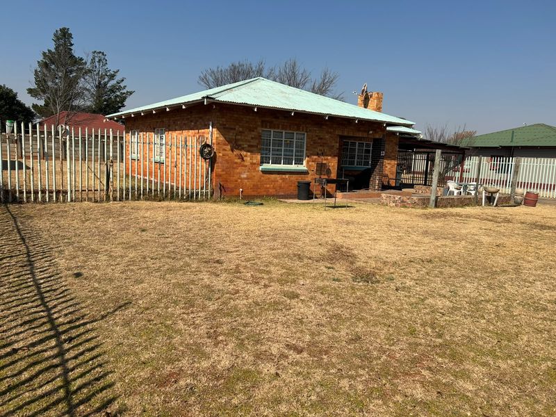 Rear view of a single-story brick house featuring a corrugated metal roof, chimney, and a paved patio area set within a fenced yard.