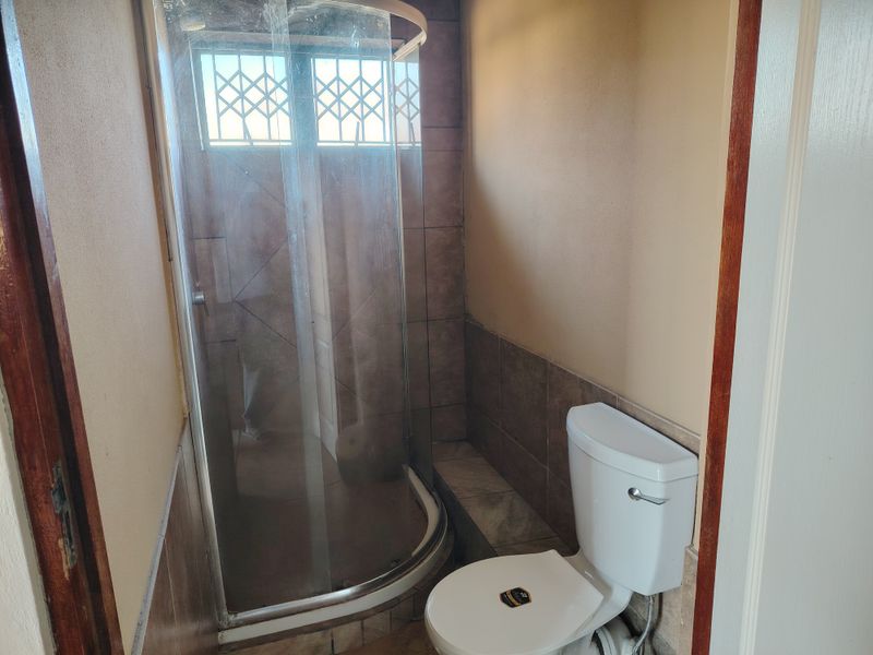 A compact bathroom featuring a curved glass shower enclosure, ceramic tiling, and a standard toilet with security bars on the window.