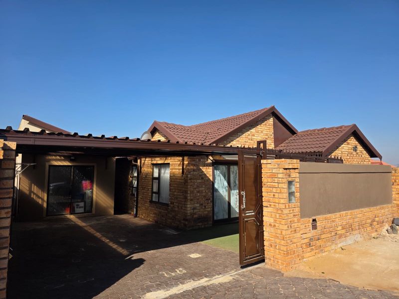 Front view of a face brick house featuring a tiled roof, paved driveway, and a covered carport with secure boundary wall and gate.