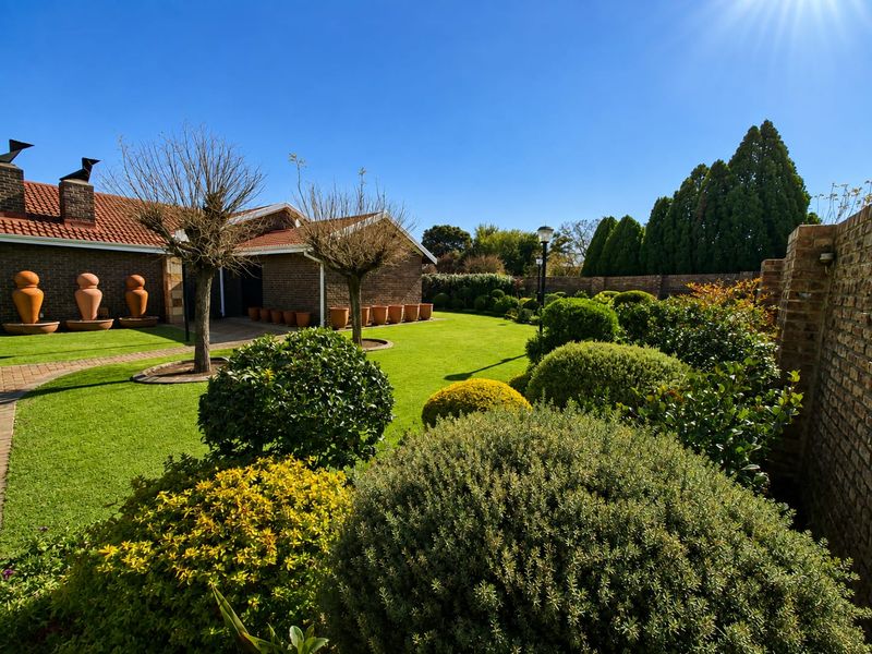 A beautifully landscaped garden featuring a lush green lawn, manicured shrubs, mature trees, and a brick-built exterior facade with a tiled roof.