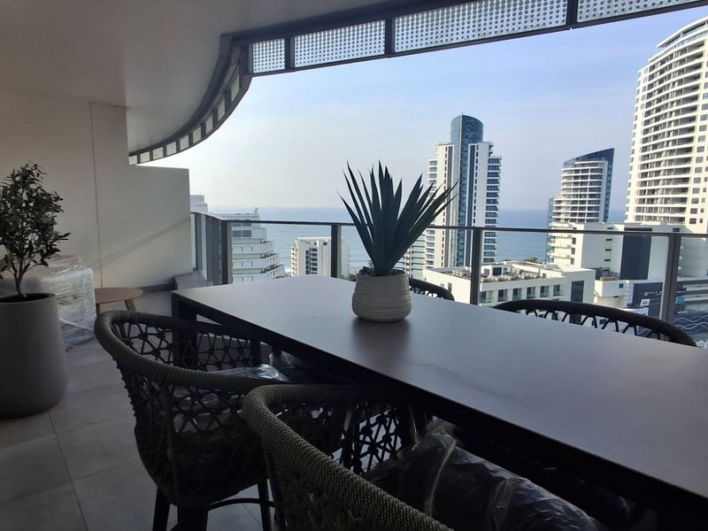 A spacious covered balcony featuring tiled flooring and a modern glass balustrade, offering expansive urban and coastal views.