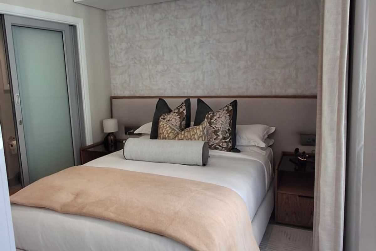 A well-appointed bedroom featuring a built-in headboard, neutral textured wallpaper, and a sleek frosted glass door leading to an en-suite bathroom.