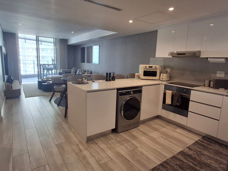 Modern open-plan apartment featuring a sleek kitchen with integrated appliances, tiled flooring, and seamless flow to a bright living area with balcony access.