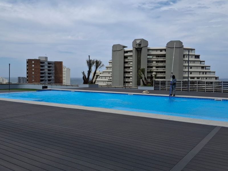 A rooftop swimming pool area featuring modern composite decking and metal safety railings with panoramic urban and coastal views.
