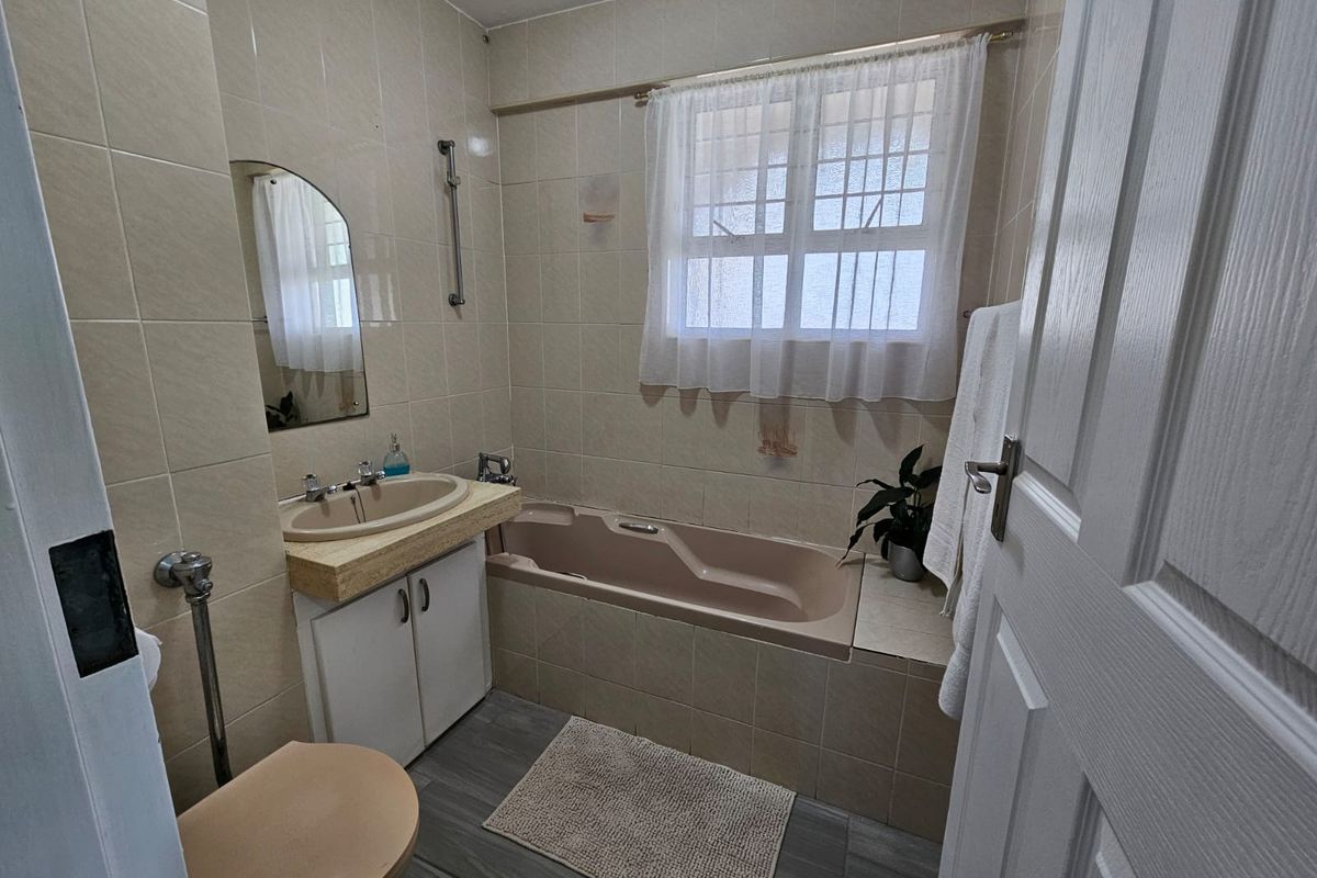 A functional bathroom featuring a built-in bathtub, vanity cabinet with basin, and neutral wall tiling, offering a dated but well-maintained space.