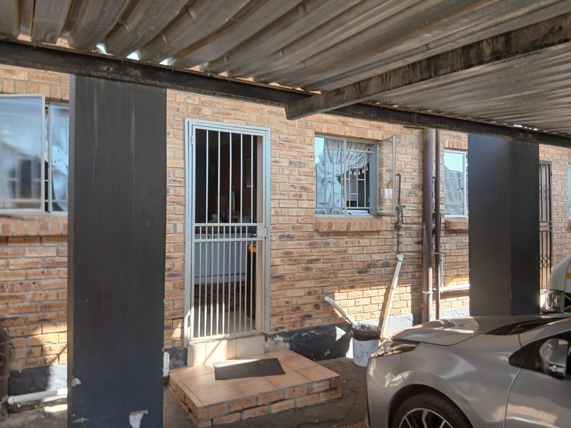 Front entrance of a townhouse featuring a brick facade, security gate, and a corrugated metal carport structure.