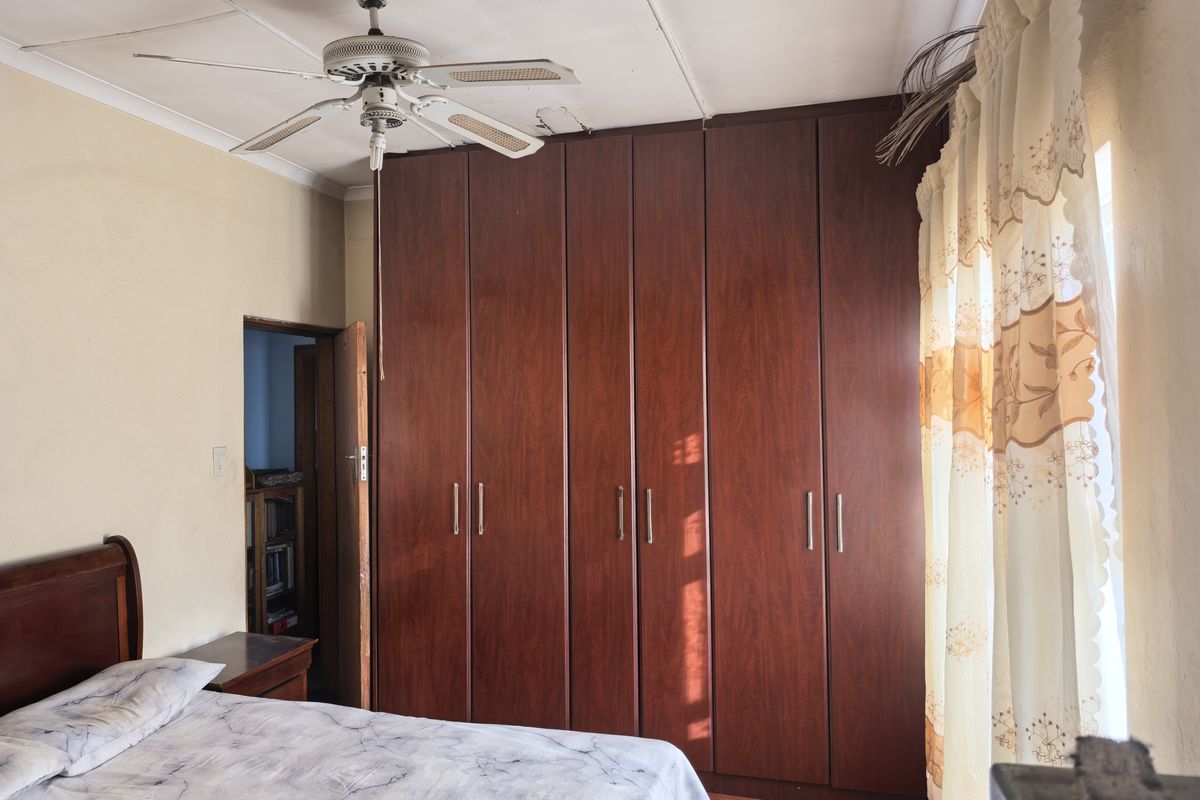 A bedroom featuring a large built-in wooden wardrobe, a ceiling fan, and carpeted flooring, showing signs of wear on the ceiling.