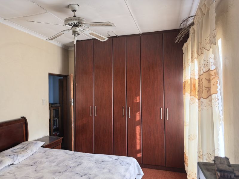 A bedroom featuring a large built-in wooden wardrobe, a ceiling fan, and carpeted flooring, showing signs of wear on the ceiling.