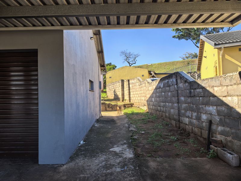 A side exterior view showing a concrete pathway, boundary wall, and covered carport area with access to the rear yard.