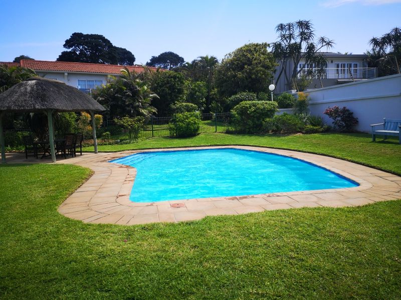 A well-maintained outdoor pool area featuring a swimming pool, paved surround, and a thatched gazebo set within a landscaped garden.