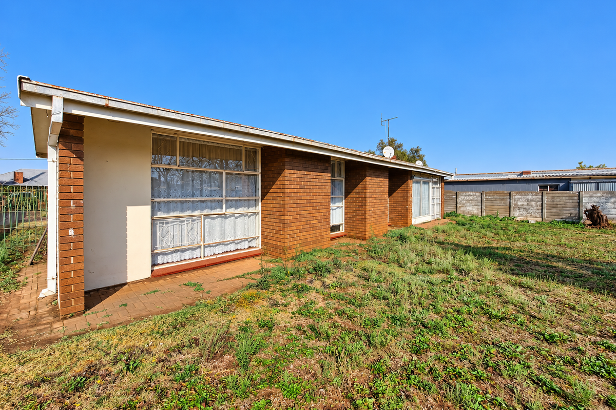 Rear view of a single-story brick house featuring a paved patio area and large windows, set within a spacious, unmaintained garden.