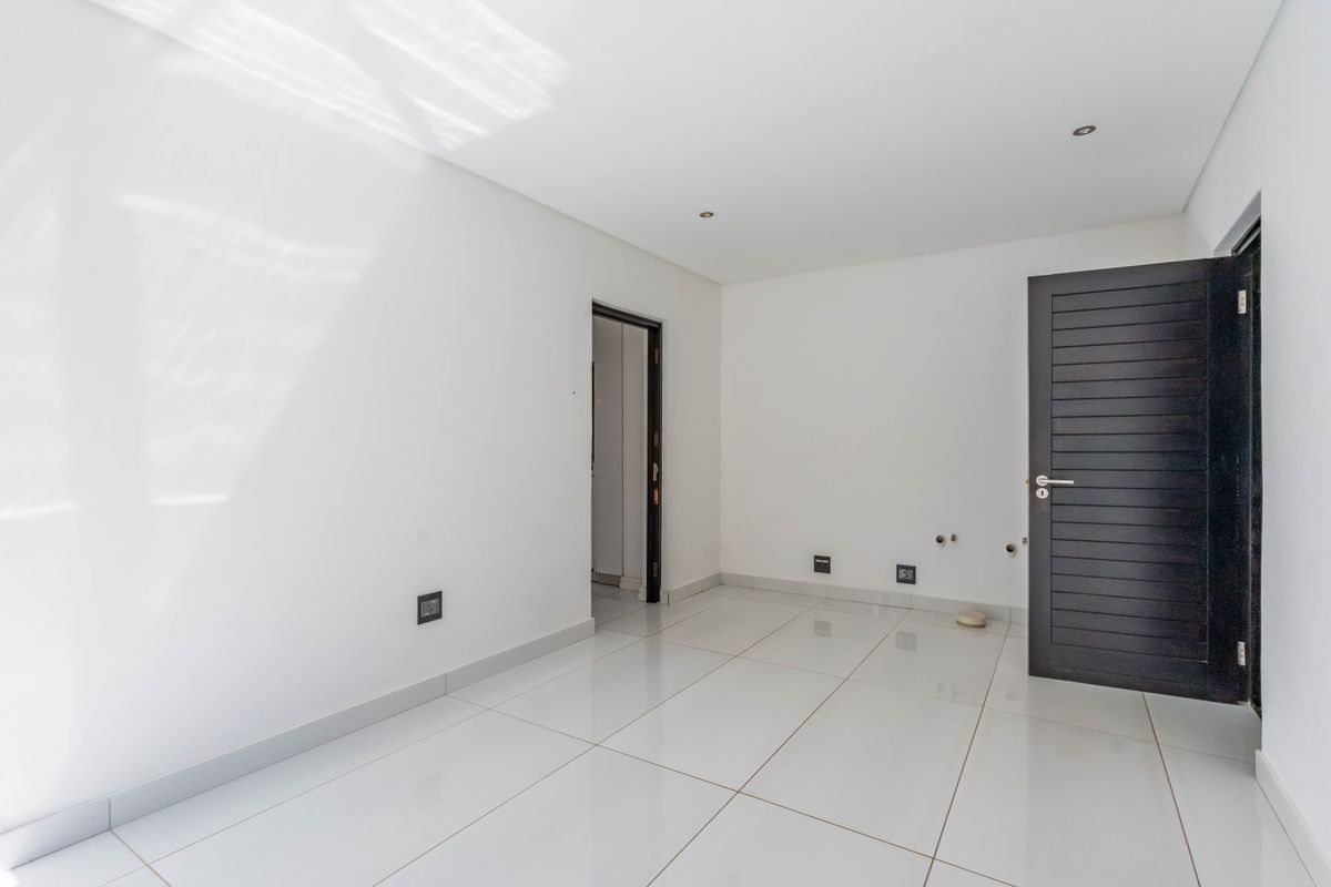 A bright, empty bedroom featuring modern white tiled flooring, recessed downlights, and a dark horizontal-slatted door.