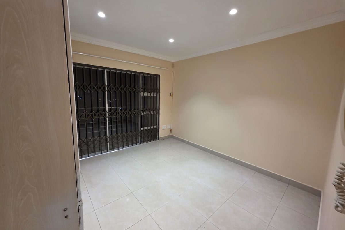 An empty, well-lit living area featuring neutral tiled flooring, recessed downlights, and a sliding glass door secured with a metal gate.