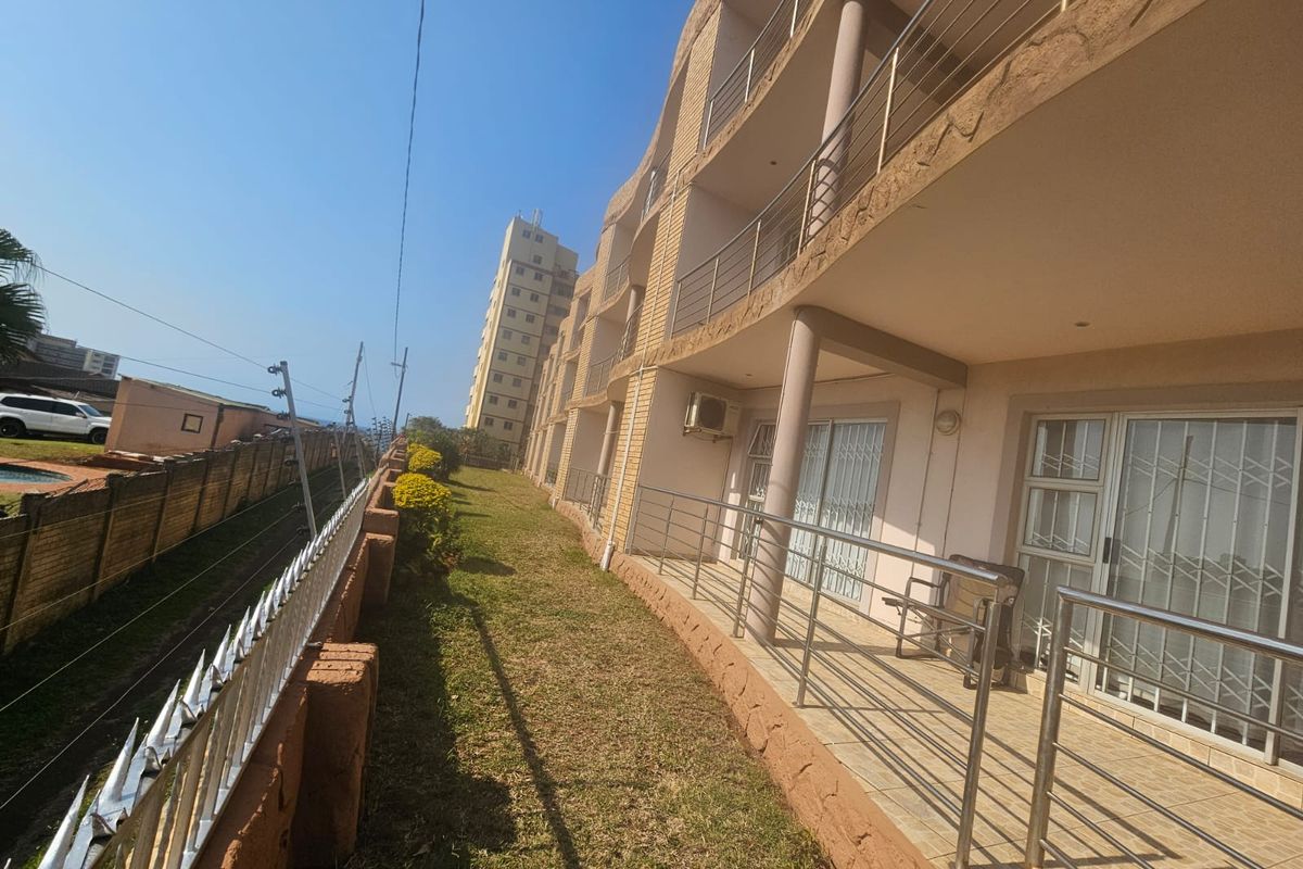 Rear exterior view of the apartment building featuring a row of balconies with metal railings, sliding doors, and security gates overlooking a grassed area.