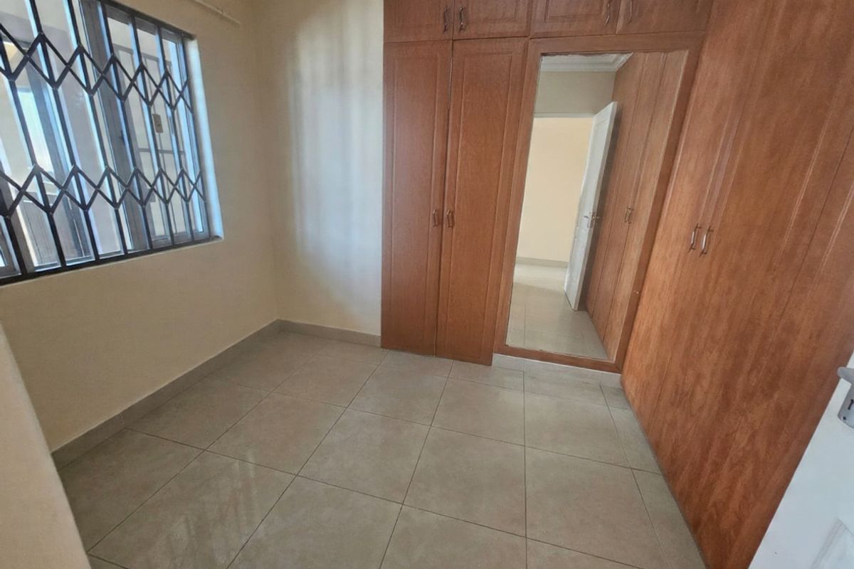 A clean, empty bedroom featuring neutral tiled flooring, built-in wooden cupboards with a central mirror, and a window fitted with a security gate.
