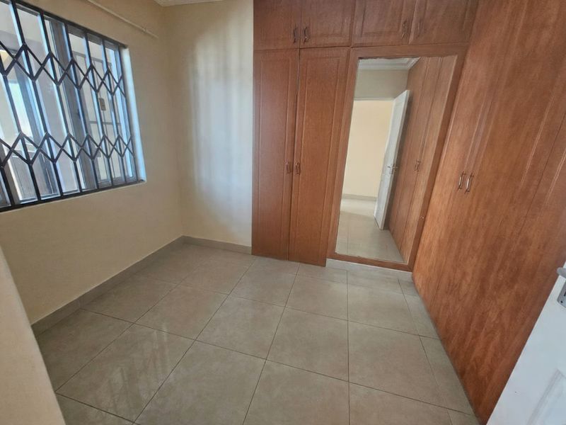 A clean, empty bedroom featuring neutral tiled flooring, built-in wooden cupboards with a central mirror, and a window fitted with a security gate.