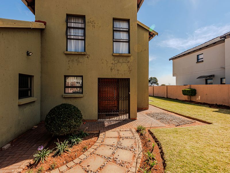 Rear view of the property featuring a paved walkway, lawn, boundary wall, and a secure entrance door with a metal security gate.