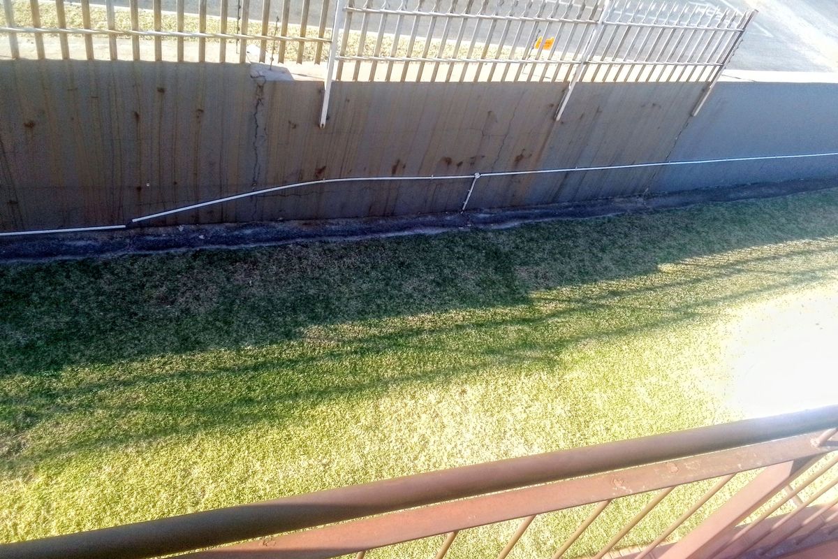 An elevated view of a rear garden area featuring a lawn, metal perimeter fencing, and a retaining wall showing signs of weathering and rust.