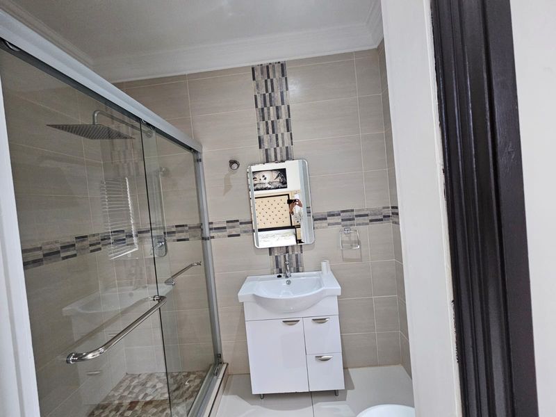 Modern bathroom featuring a glass-enclosed shower with a rain shower head, a white vanity cabinet with a basin, and neutral wall tiling with mosaic accents.