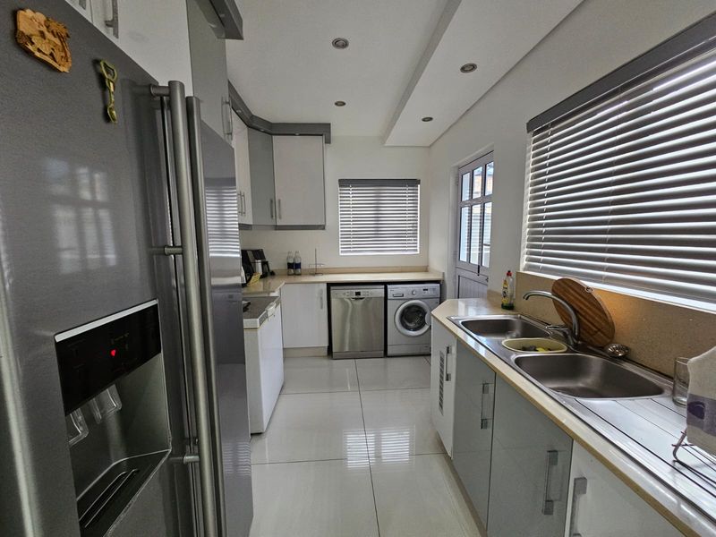 A functional scullery featuring modern cabinetry, double stainless steel sink, tiled flooring, and integrated laundry appliances with direct access to the exterior.